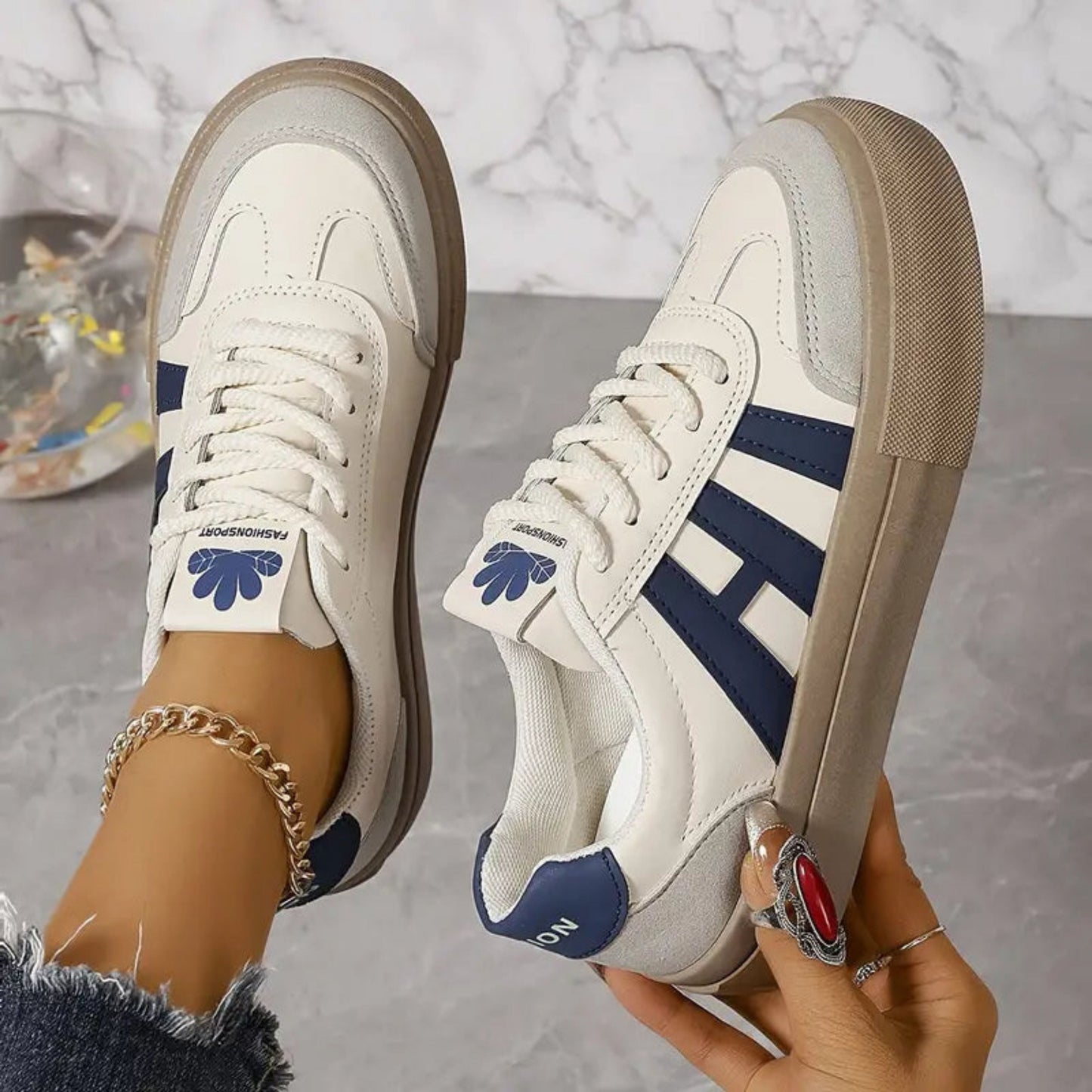 White sneakers with blue 3-stripe detail, lace-up, tan sole, floral patch on tongue.
